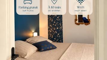 A bedroom with a bed, pillows, a nightstand, a wall-mounted lamp, a decorative mirror, and a wall with icons indicating parking, a location marker, and Wi-Fi availability.