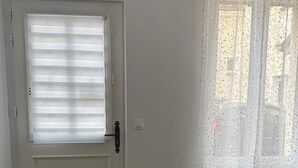 Ett rum med en vit dörr, ett fönster med tunna, genomskinliga gardiner, ett runt bord och en blå stol.