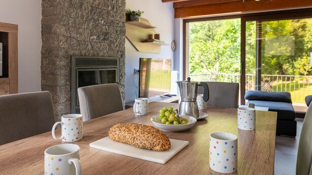 Un comedor con pared de piedra, chimenea, una mesa con un pan, un bol de uvas y una cafetera.