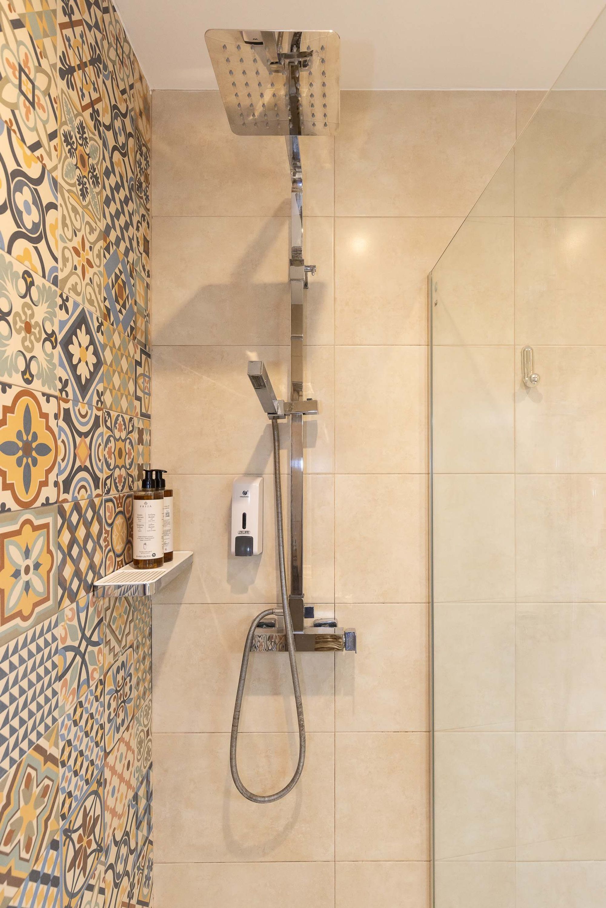 Una ducha con manguera de mano y cabezal de ducha en la pared, un revestimiento de azulejos con diseño geométrico y un estante pequeño con artículos de aseo.