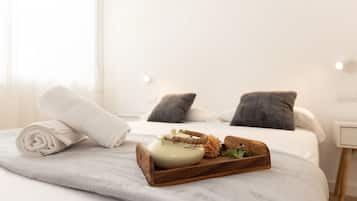 Una cama bien hecha con una bandeja de madera que sostiene una tetera, flores, una toalla enrollada y una almohada sobre un cubrecolcha blanco.