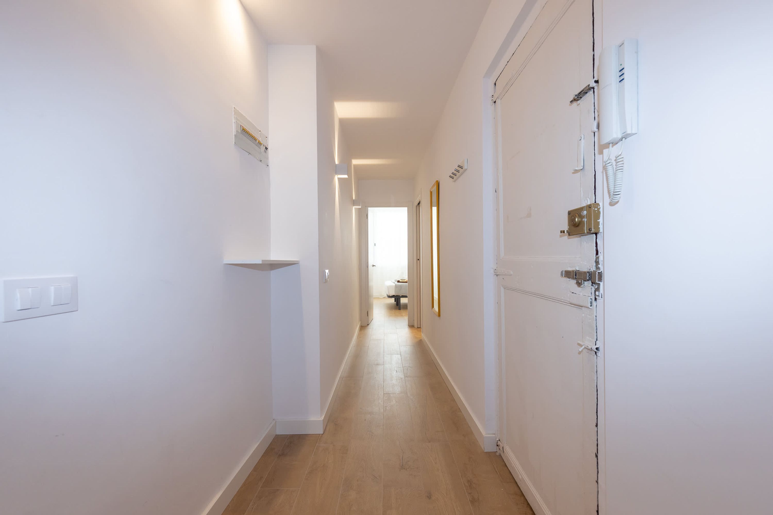 A narrow hallway with white walls, a wooden floor, and a door with a lock and a phone on the wall.