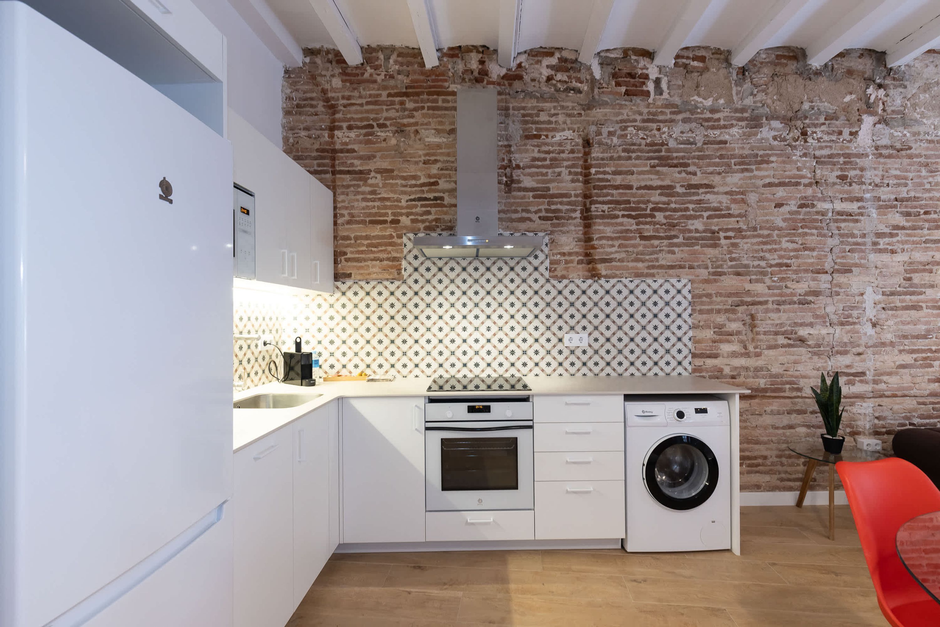 A compact kitchen with a brick wall, a washing machine, and a red chair.