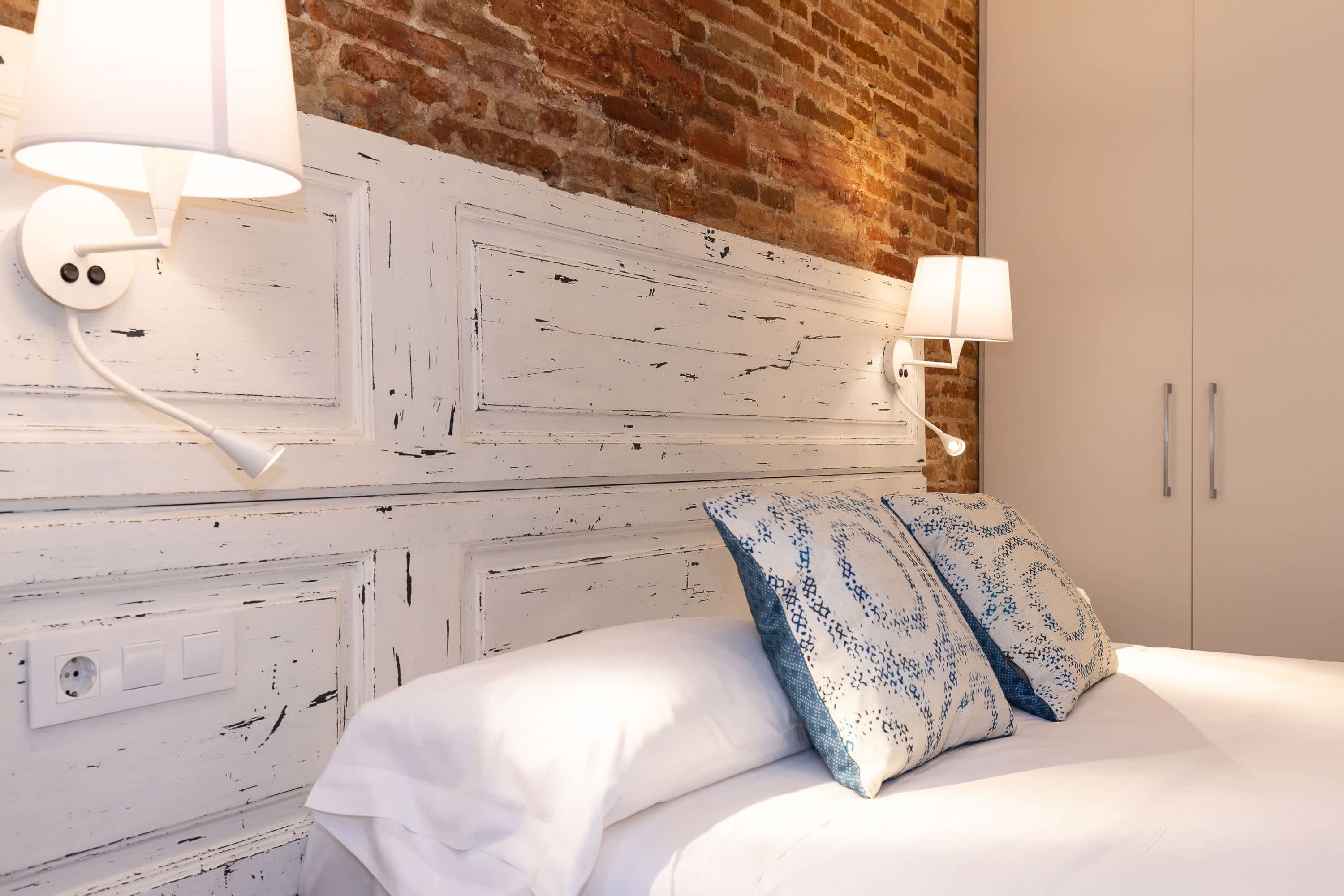 Un dormitorio con cama blanca, almohadones estampados, cabecera blanca con pintura desgastada y dos lámparas fijadas a la pared.