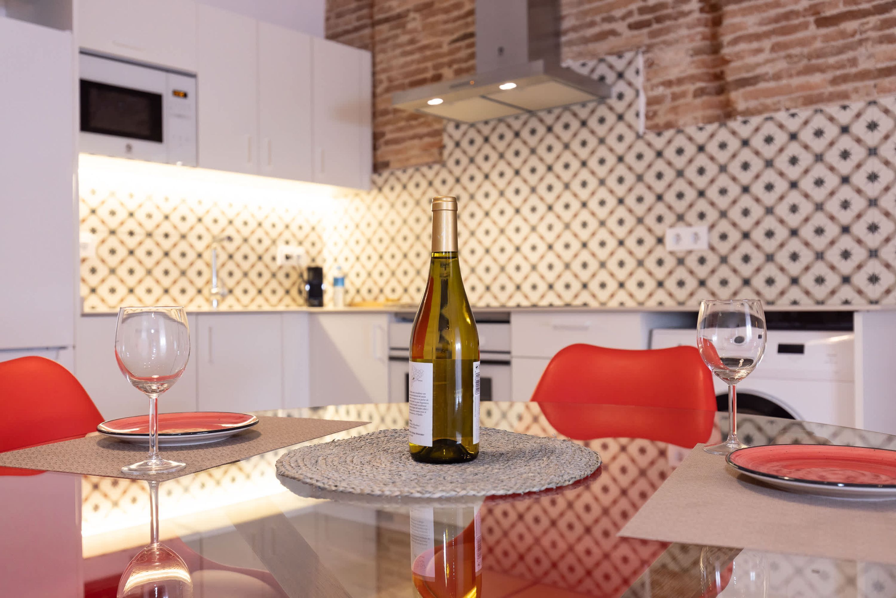 Een eettafel met een wijnfles, twee glazen en rode borden in een moderne keuken met witte kasten en een decoratieve achterwand.