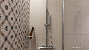 Una ducha con cabezal de ducha manual, mampara de vidrio y un dispensador de papel higiénico en la pared.