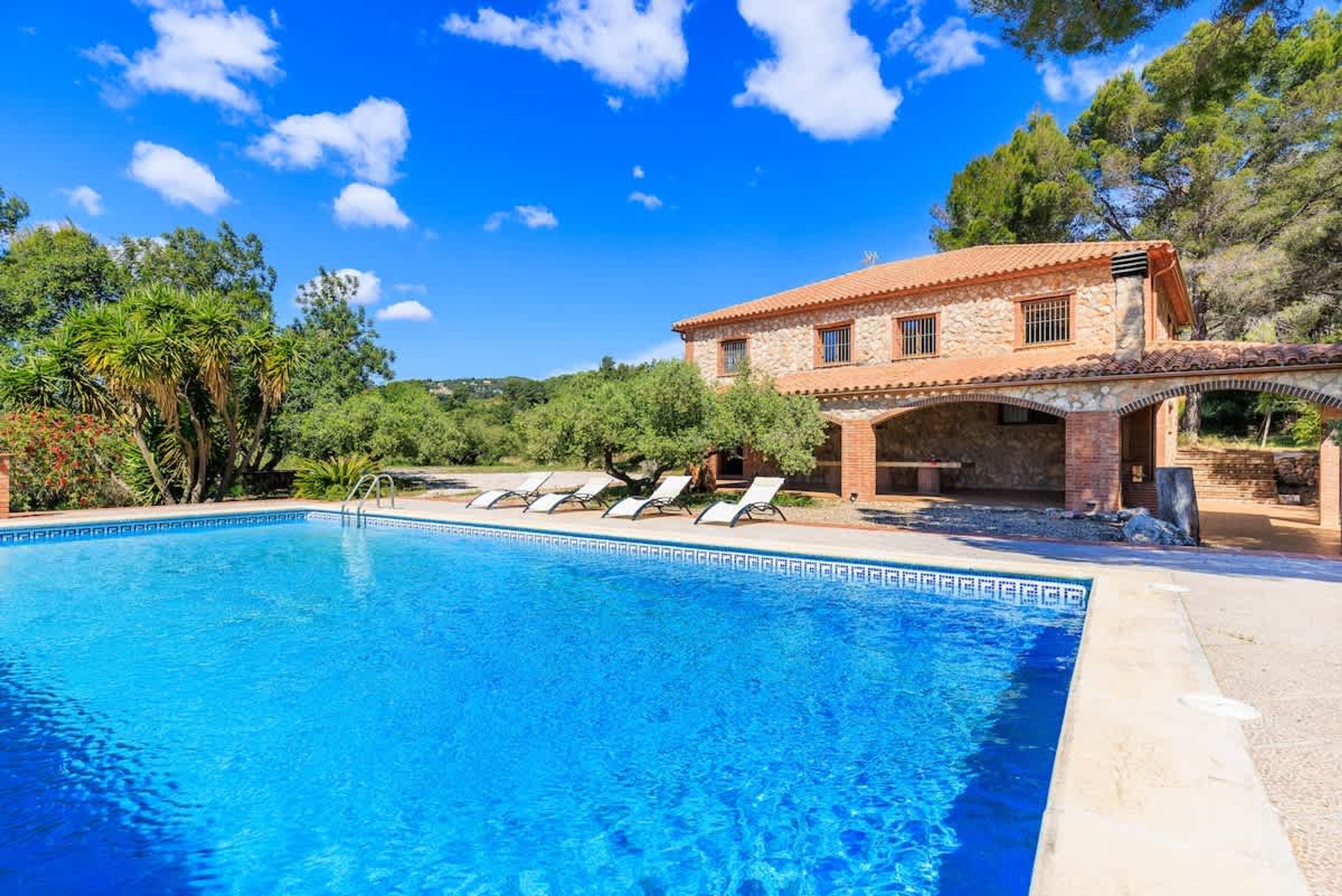 Una piscina con sillas de descanso, un edificio de piedra con techo de tejas y follaje circundante.