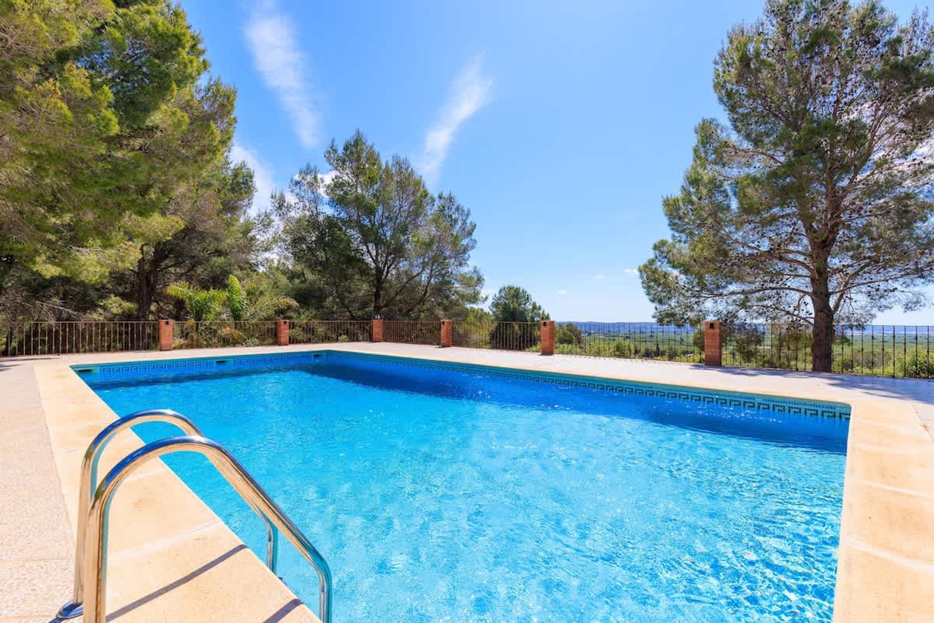 Una piscina rectangular con una escalera metálica, rodeada por un solado de baldosas y una cerca de metal.