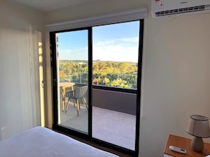 A hotel room with a large sliding glass door leading to an outdoor balcony, a bed, a table with chairs, and a lamp on a nightstand.