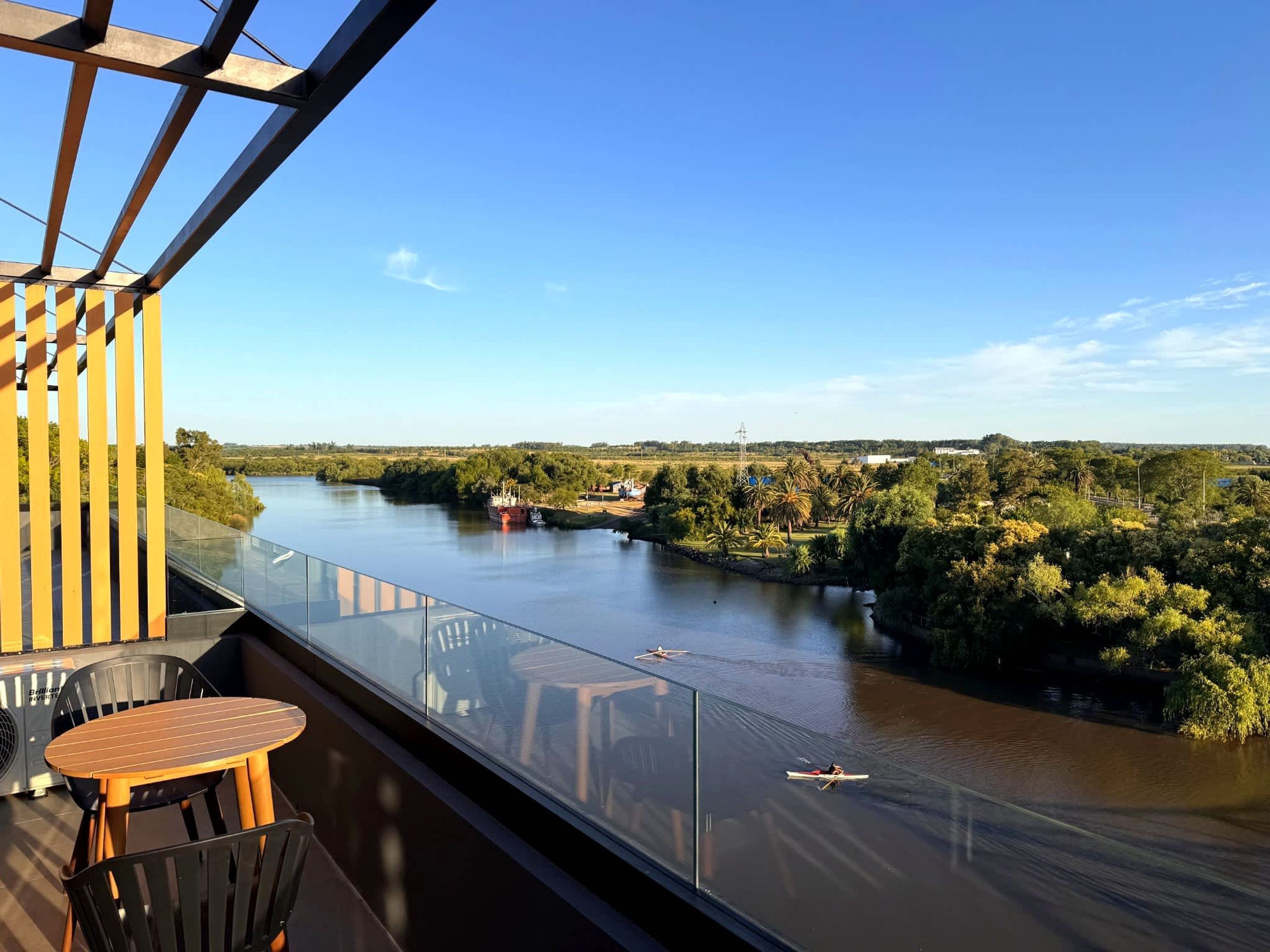 A balcony with a glass railing overlooking a river, a wooden table, and chairs. - Carmelo Postal in Carmelo (Carmelo)