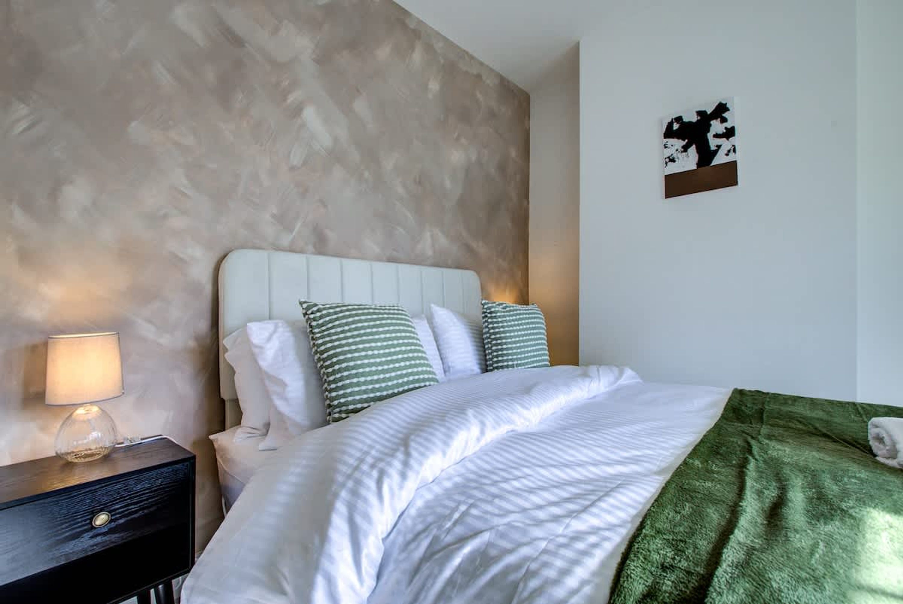 Une chambre à coucher avec un lit, des oreillers, une table de chevet, une lampe et une œuvre d’art murale.