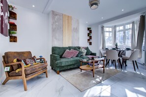 A living room with a green sofa, wooden armchairs, a coffee table, and a dining area with a table and chairs. - Perfect Booking Stylish 3 Bed House in Enfield (Enfield)