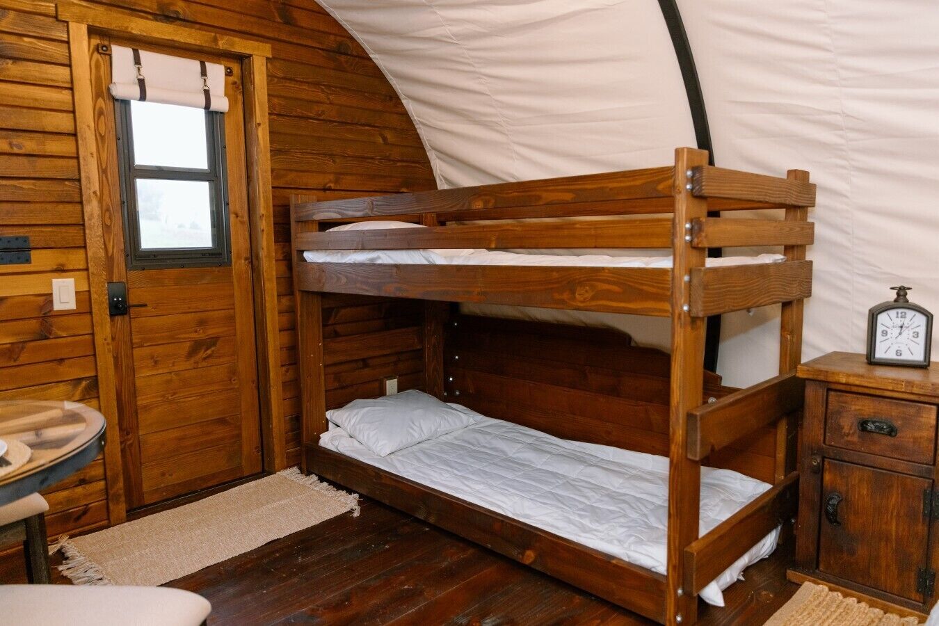 Un lit superposé dans une pièce aménagée comme une tente, avec une porte en bois, une petite table et une horloge posée sur un meuble en bois.