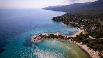 An aerial view of a coastal area with clear waters, a rocky outcrop, and a beach with buildings and greenery.