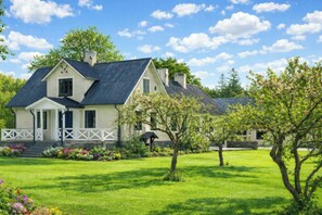 A two-story house with a front porch, a well-maintained lawn, and flowering trees. - Farm on Gotland with fruit trees near harbour (Slite)
