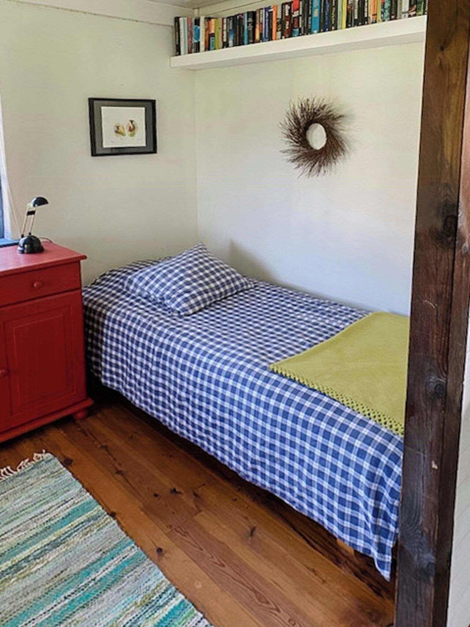 Une petite chambre à coucher avec un lit, une commode rouge, une bibliothèque et un tableau encadré au mur.