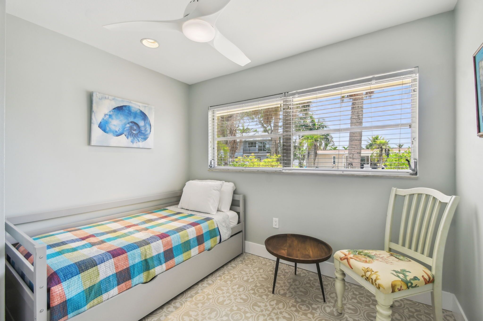 A small, well-lit room with a single bed, a chair, a window with blinds, and a ceiling fan.