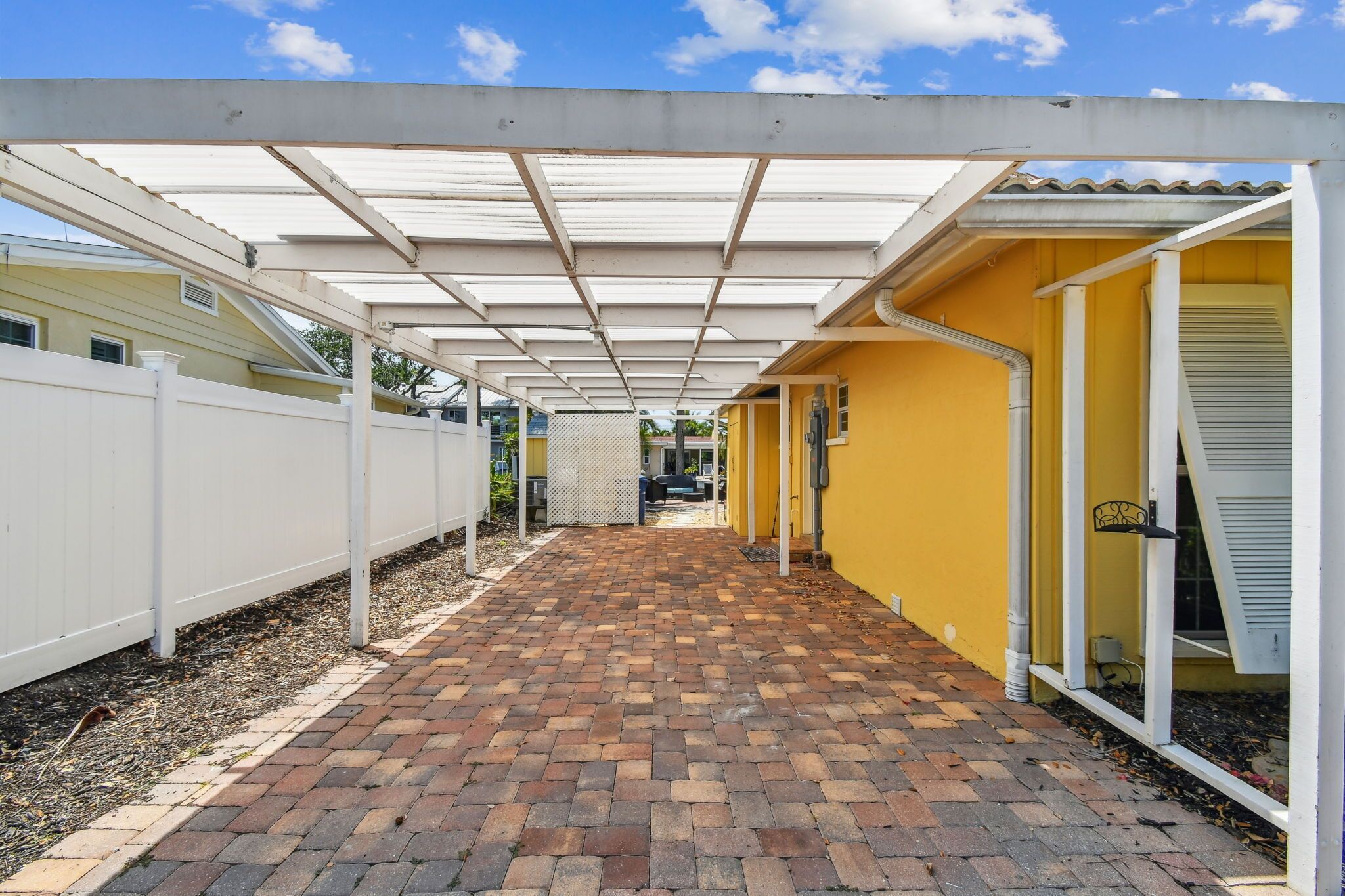 Une terrasse couverte, avec un chemin en briques menant à un bâtiment jaune, délimité par une clôture blanche et doté d’une porte blanche.