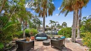 An outdoor patio with wicker furniture, potted plants, and a view of a building in the background. - Sunnyside Up Cottage - Walk to Siesta Beach, Waterfront Home by The Bay & Key Collection (Siesta Key)
