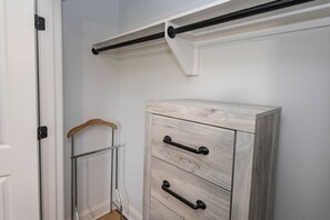 A white wardrobe with three drawers and black handles, a wooden hanger, and a metal rack against a white wall.