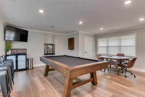 A pool table in a room with a television above it, a cabinet, and a dining area with chairs. - Pines at 5 Corporate Furnished Rentals Suite 214 (Hephzibah)