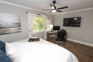 A bedroom with a bed, a desk with a computer, a chair, a ceiling fan, and a TV on the wall.