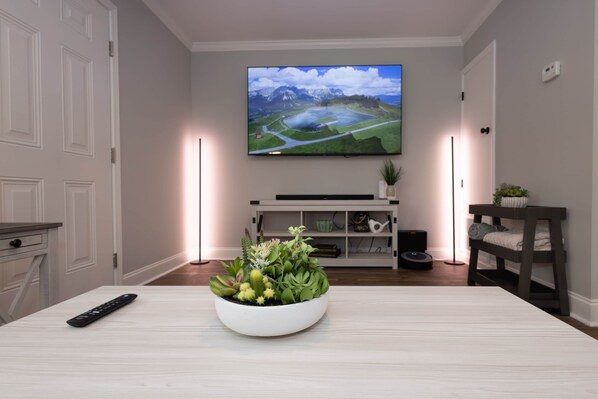 A living room with a flat-screen TV mounted on the wall, a white console table, a remote control, a plant, and a bench.