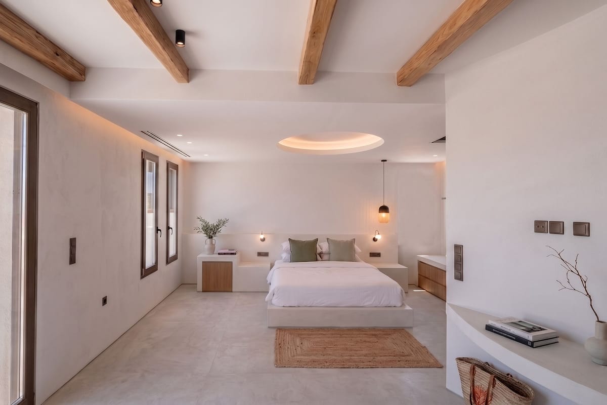A bedroom with a large bed, bedside tables, a nightstand, a wall-mounted lamp, and a view of the outdoors.