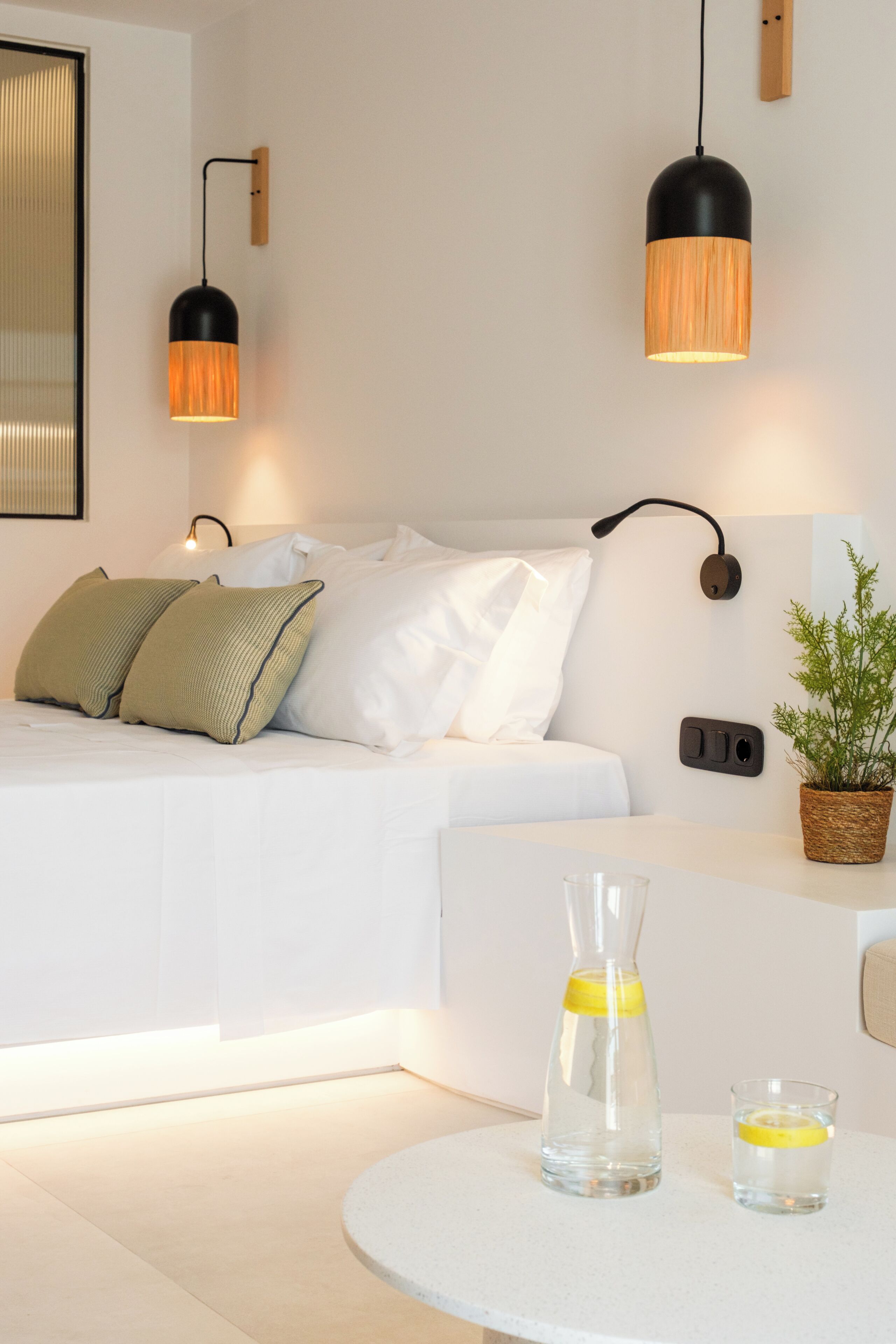 Ein modernes Schlafzimmer mit einem Bett, Nachttischlampen, einer Topfpflanze und einem runden Tisch mit einem Glasbehälter und einem Glas.