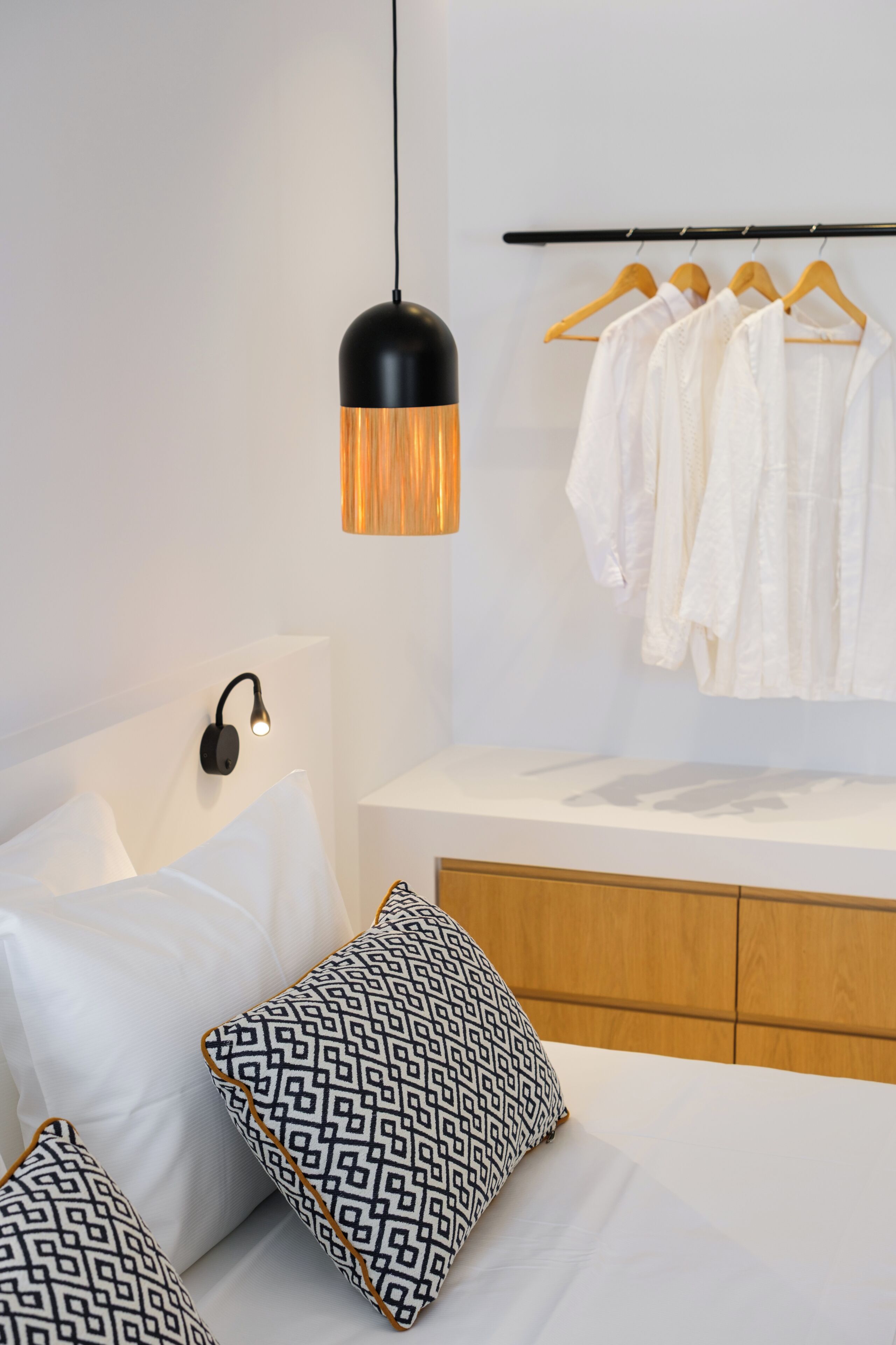 Quarto moderno com cama, travesseiros, luminária pendente, mesa de cabeceira e um cabideiro.