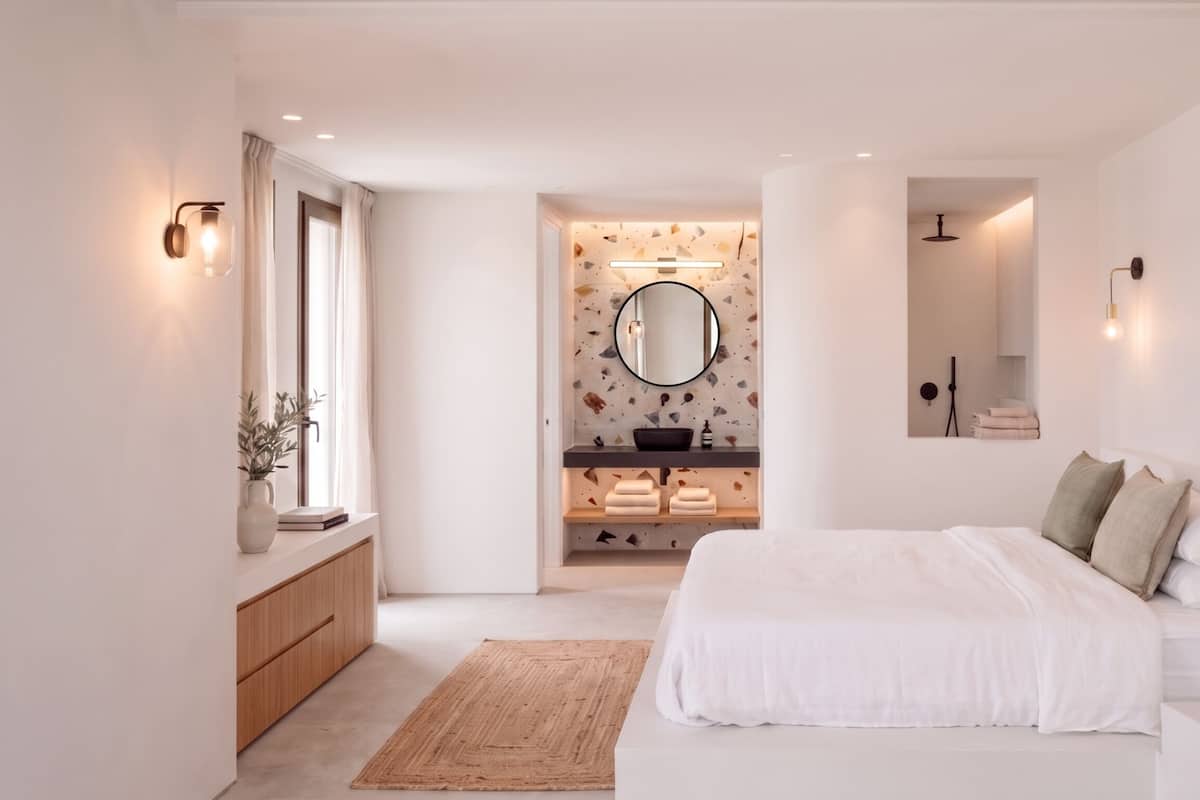 A modern bedroom with a bed, bedside tables, a mirror, and a bathroom with a sink and towel rack.