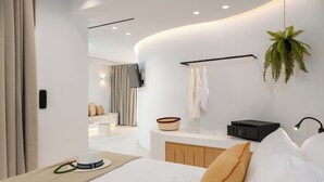 A modern bedroom with a curved wall, a bedside table, a bed, a hanging plant, and a TV mounted on the wall. - The Lofts Paros Executive Suites (Naousa)