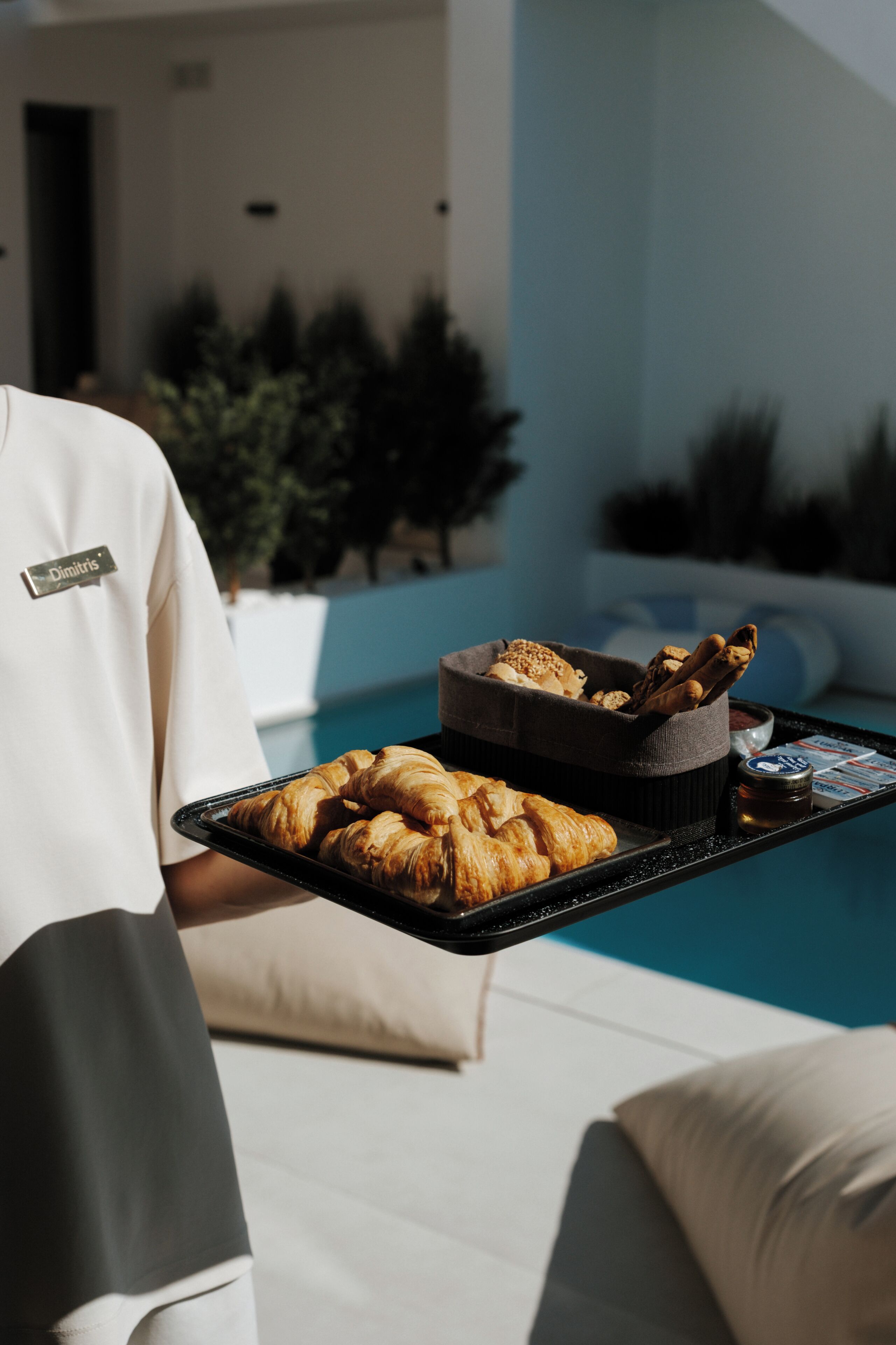 Una bandeja con pasteles y un cuenco pequeño, sostenida por una persona con un gafete, cerca de una piscina con un edificio moderno y vegetación al fondo.