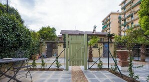 Un cortile recintato con cancello in metallo, piante in vaso, un tavolo in pietra e un edificio con balconi sullo sfondo.
