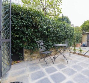 Un patio con suelo de piedra, una mesa y sillas de metal, y una pared de piedra con plantas trepadoras.