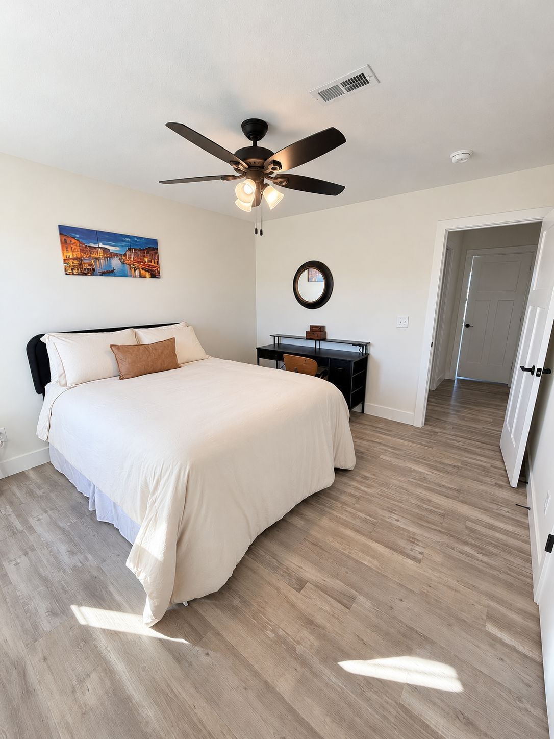 Une chambre à coucher comprenant un lit, un bureau, un ventilateur de plafond, un tableau et une porte donnant sur une autre pièce.
