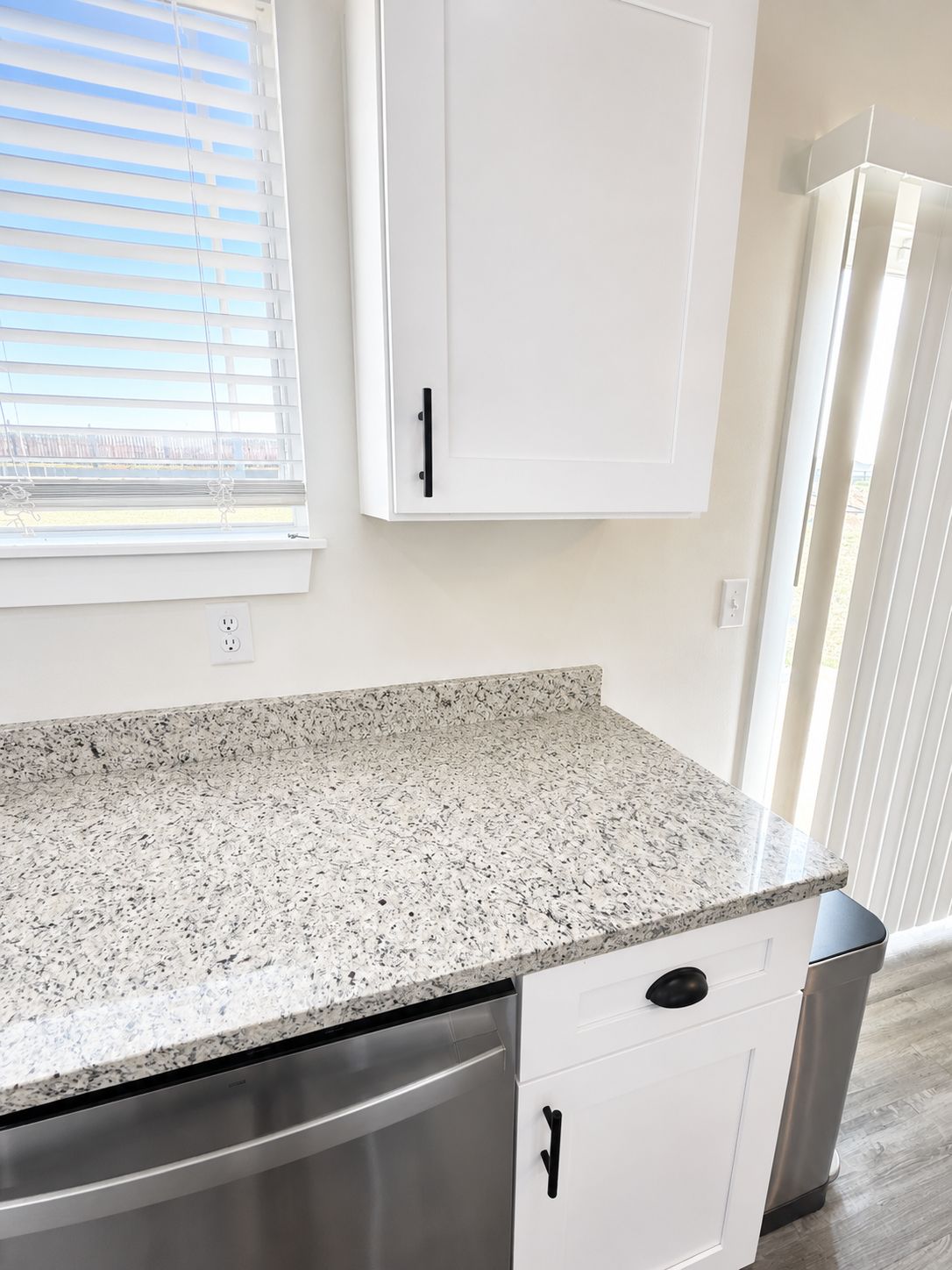 A kitchen with white cabinets, a granite countertop, a dishwasher, and a trash can.
