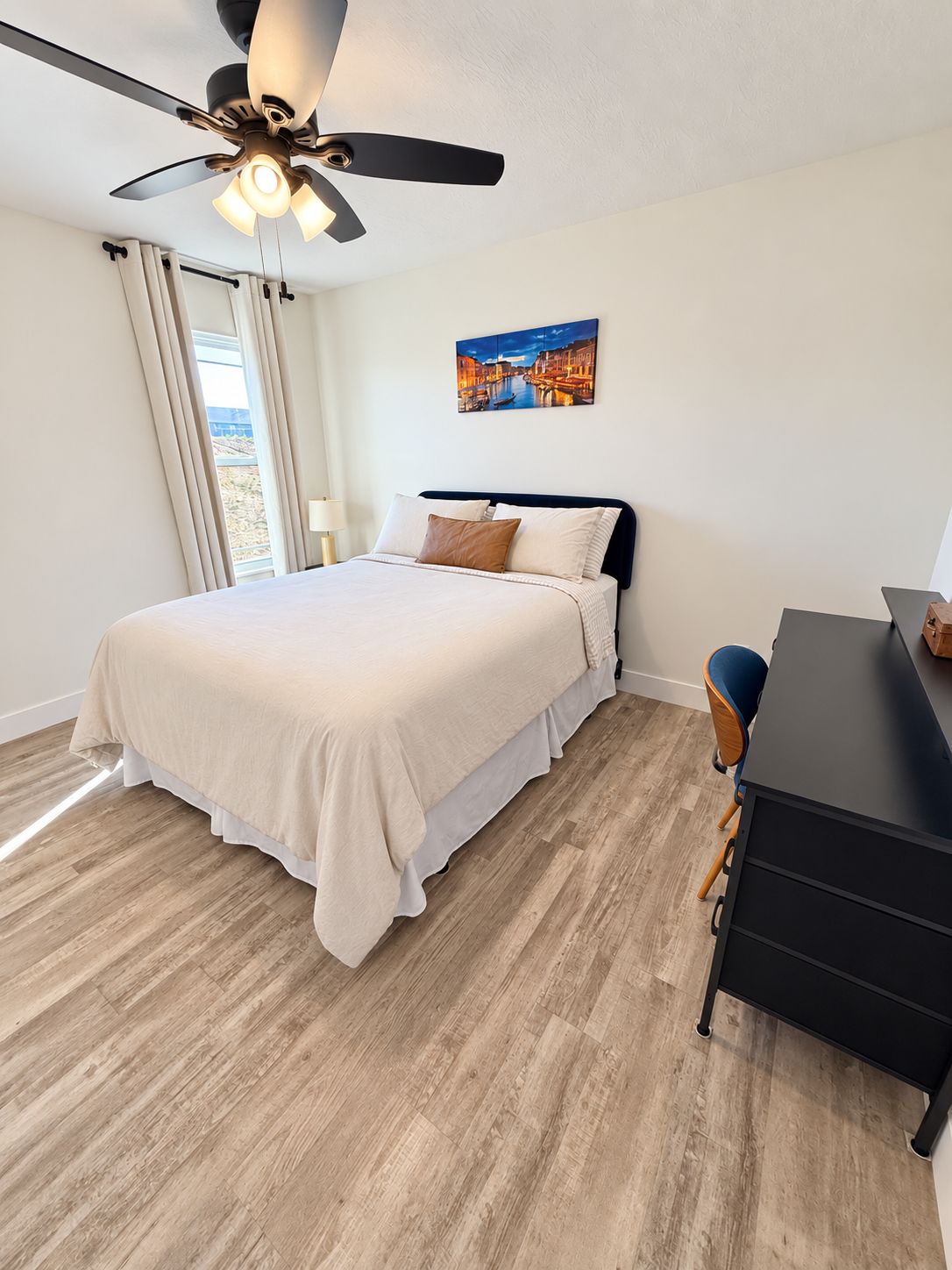 Une chambre à coucher avec un lit, un bureau, une chaise, un ventilateur de plafond et un tableau accroché au mur.