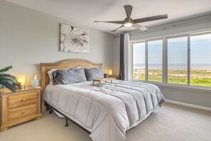 A bedroom with a bed, bedside tables, a ceiling fan, a large window with a view, and a wall-mounted artwork. - Sandy Toes: Harbor Isl Pool & Beach Retreat (Harbor Island)