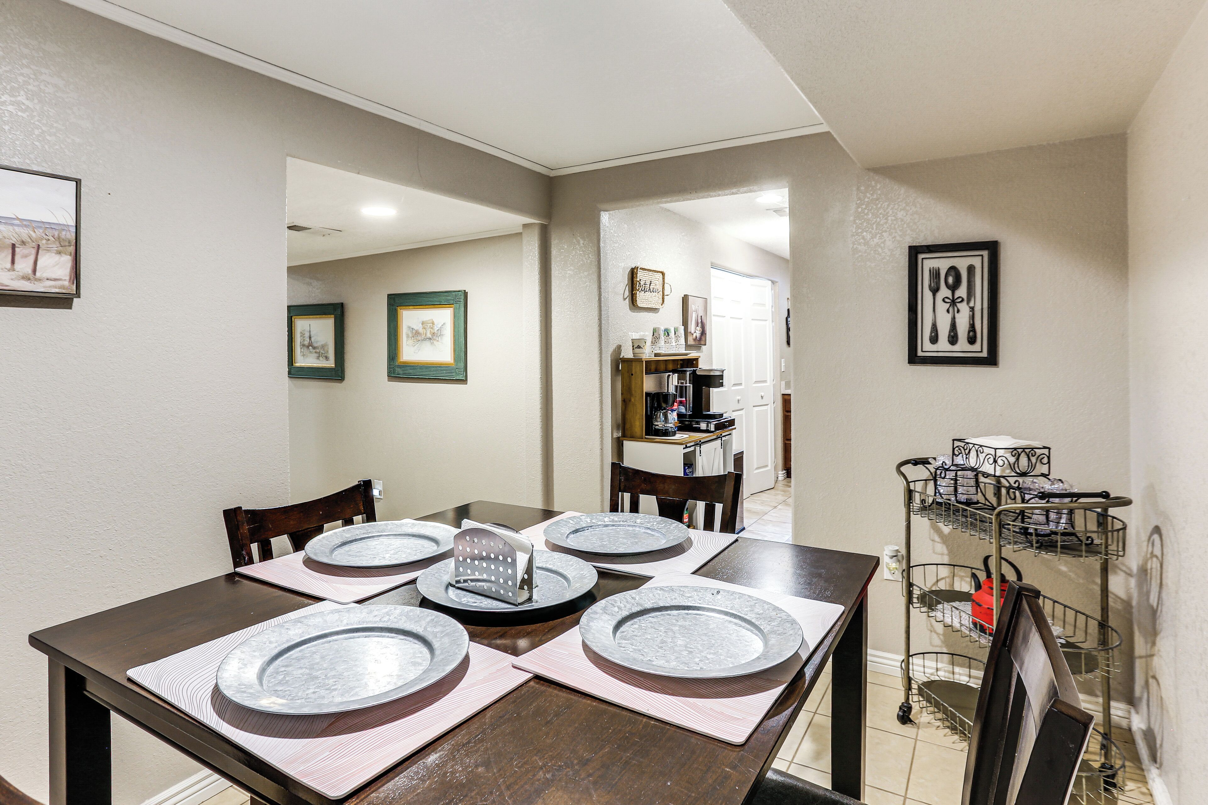 Un coin repas avec une table dressée pour quatre personnes, une cuisine en arrière-plan et un étagère en métal remplie de vaisselle.