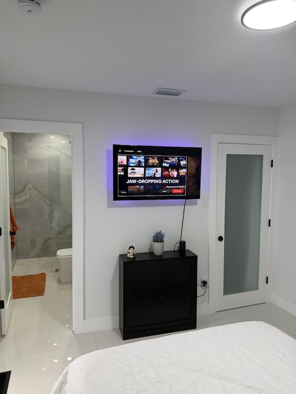 A modern bedroom with a flat-screen TV mounted on the wall, a black entertainment unit below, and a white bed in the foreground.