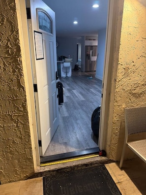A doorway leading to a room with a wooden floor, a white door with a window, and a small kitchen area visible in the background.