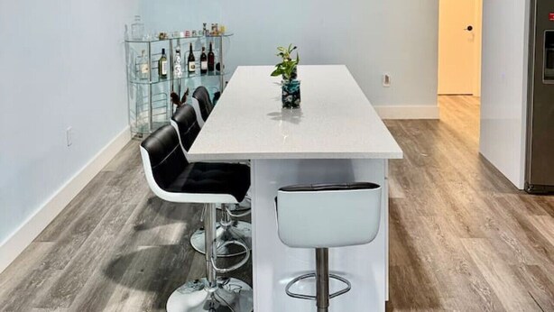 Une cuisine moderne avec une table blanche, des chaises noires, un meuble vitré contenant des bouteilles et une horloge au mur.