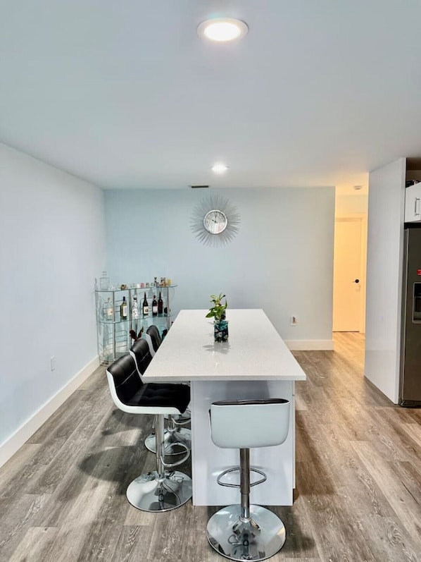 A modern kitchen with a white table, black chairs, a clock, and a glass cabinet with bottles.