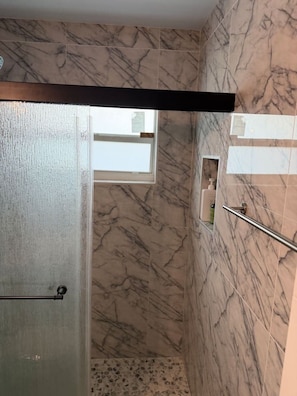 Ein Badezimmer mit MarmorwĂ€nden, einer GlasduschtĂŒr, einem Fenster und einer kleinen Ablage fĂŒr Toilettenartikel.