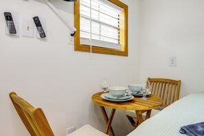 Una habitación con una mesa de madera preparada para dos personas, una ventana con persianas y dos controles remotos en la pared.