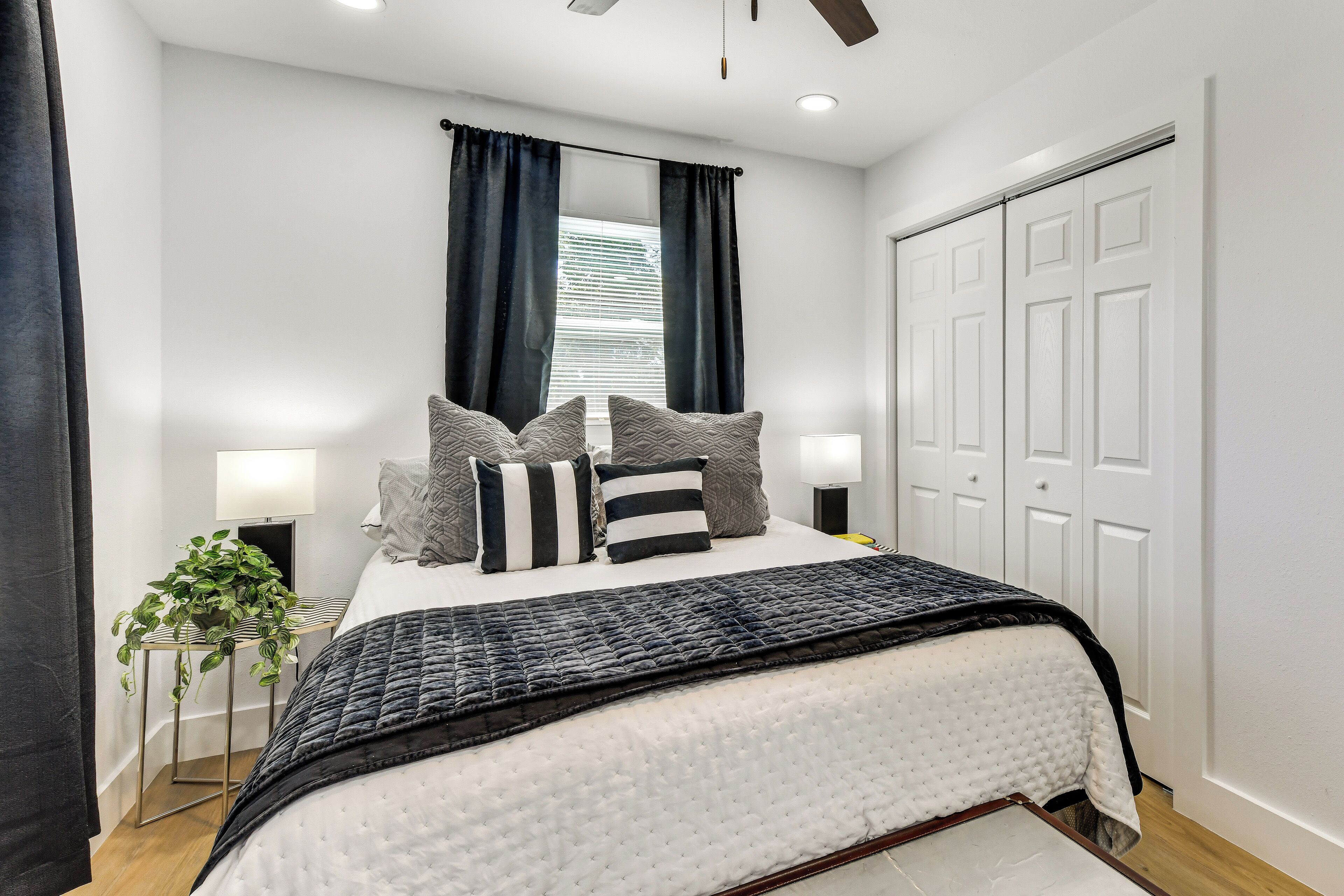A bedroom with a bed, bedside lamps, a window with curtains, a plant, and a door.