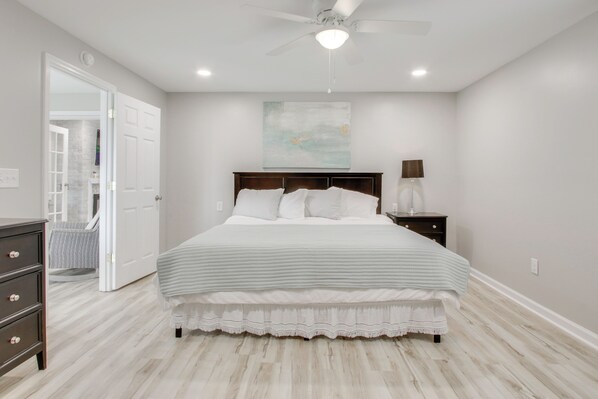 A bedroom with a bed, a nightstand, a lamp, a ceiling fan, and a door leading to another room.