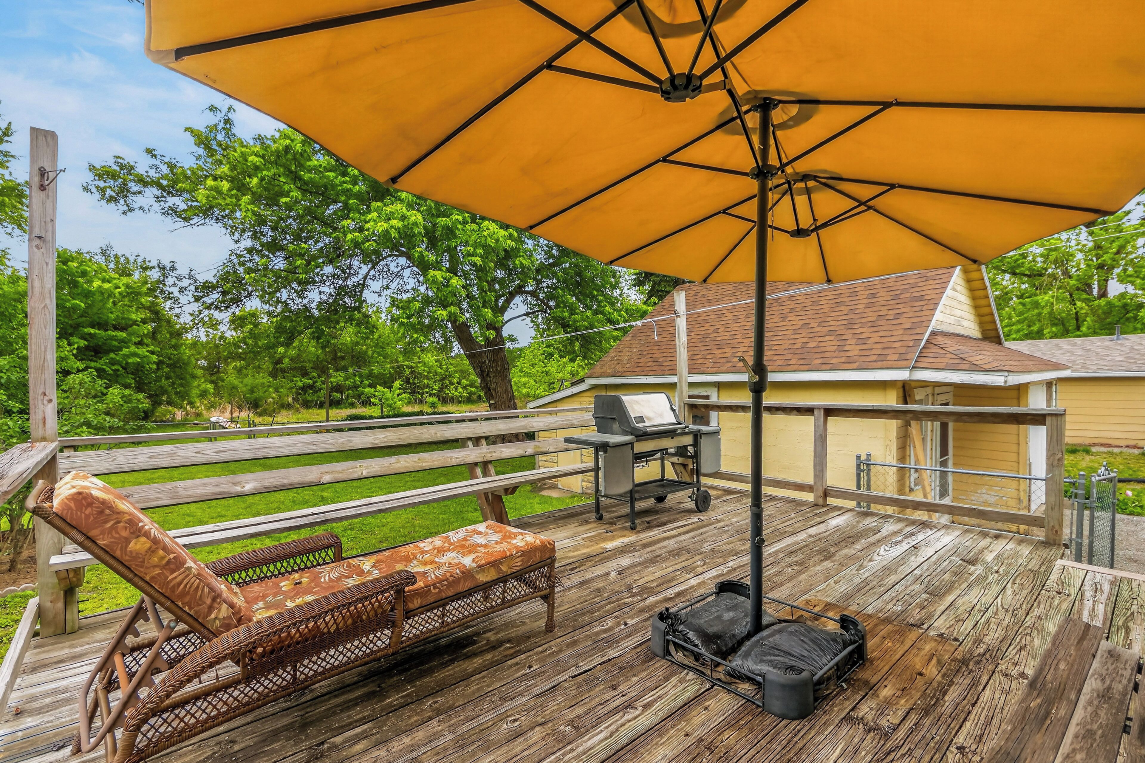 A wooden deck with a large yellow umbrella, a wicker lounge chair, a grill, and a small table.