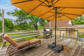Una terraza de madera con un gran paraguas amarillo, una silla de mimbre, una parrilla y una mesita.